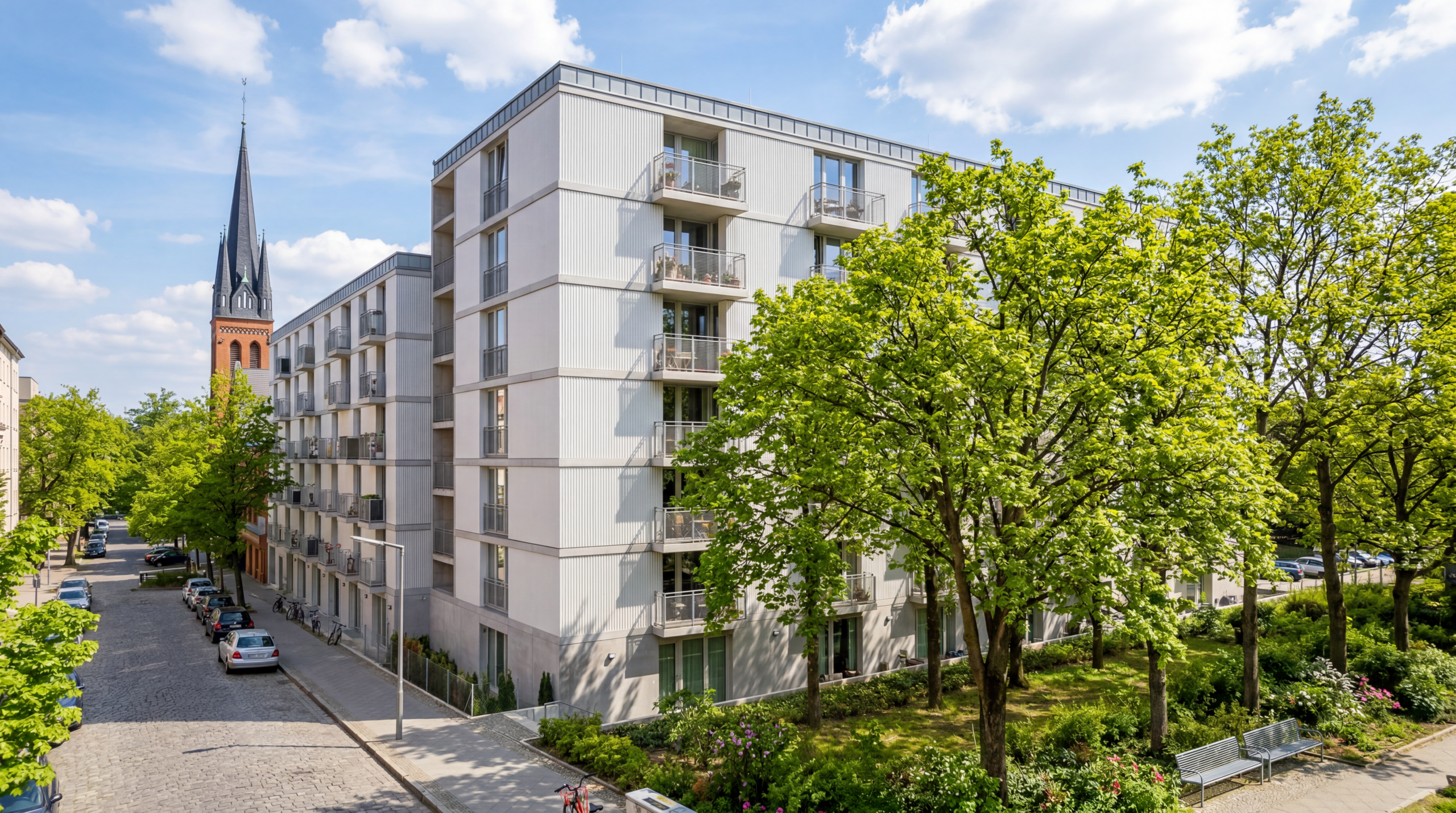 Straße mit mehrstöckigem Wohngebäude und Bäumen, im Hintergrund Kirchturm mit spitzem Dach